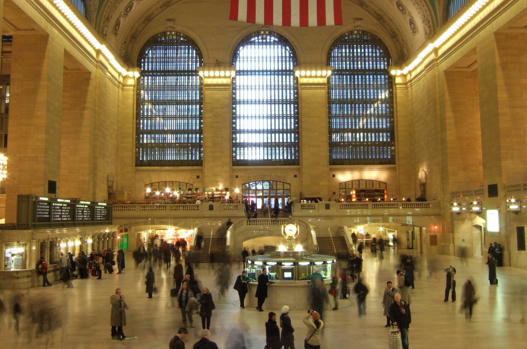 Commuters, blurred by movement, cross the main concourse of Grand Central Terminal in New York beneath three tall arched windows and a hanging American flag