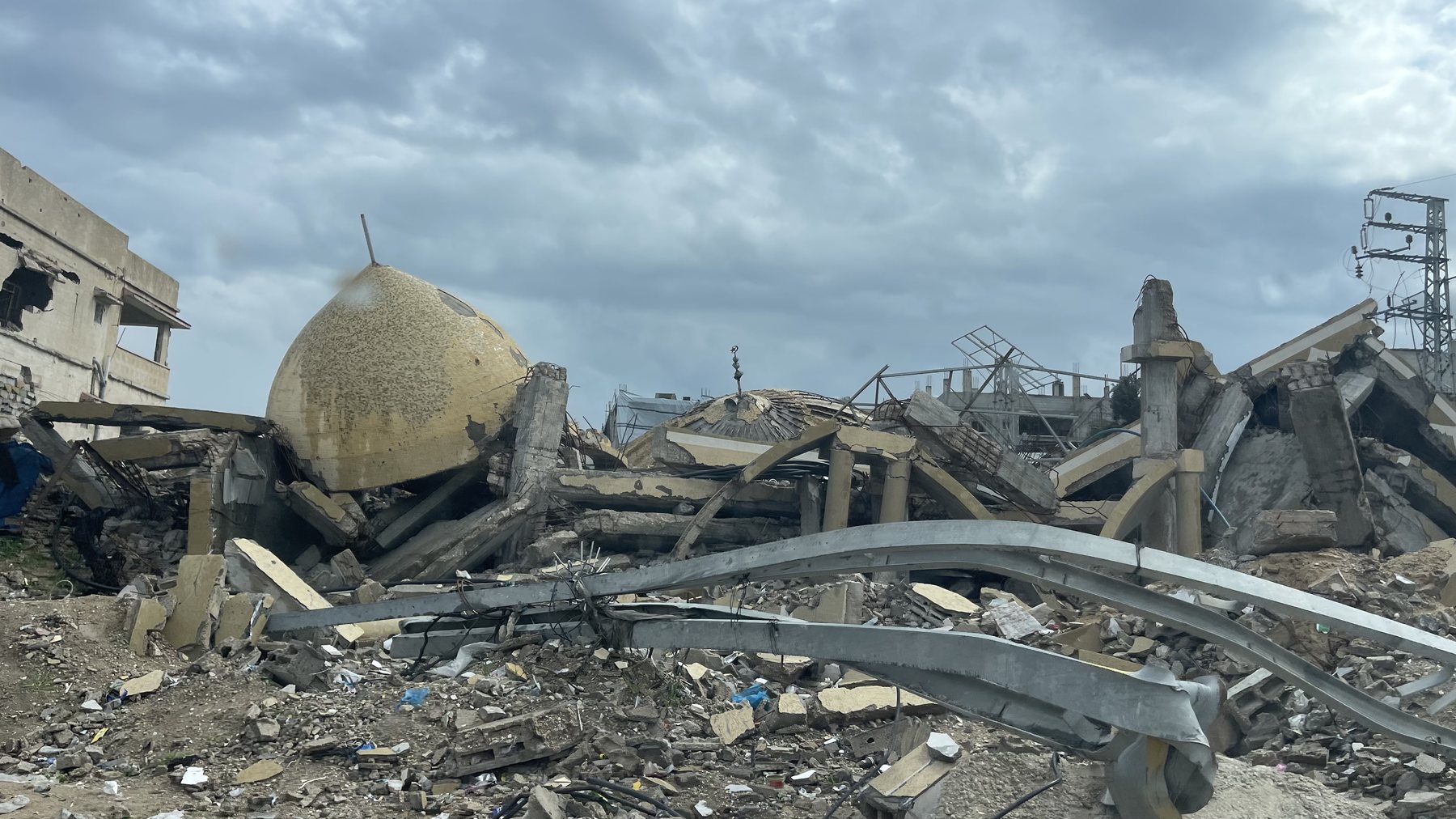 The ruins of a mosque in Jabalia: a golden dome lying tilted on a heap of broken concrete slabs and twisted steel under a grey sky, a damaged apartment block to the left
