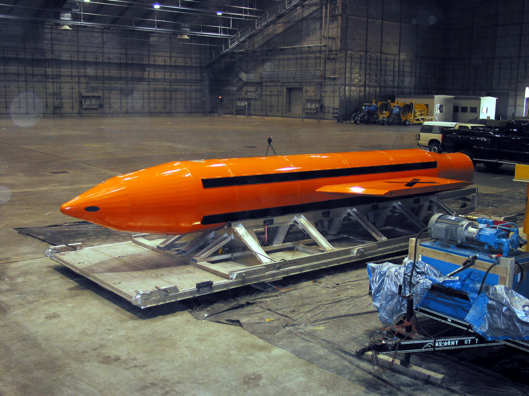 A long orange bomb with a pointed nose and small fins rests on a metal cradle on the floor of a large hangar