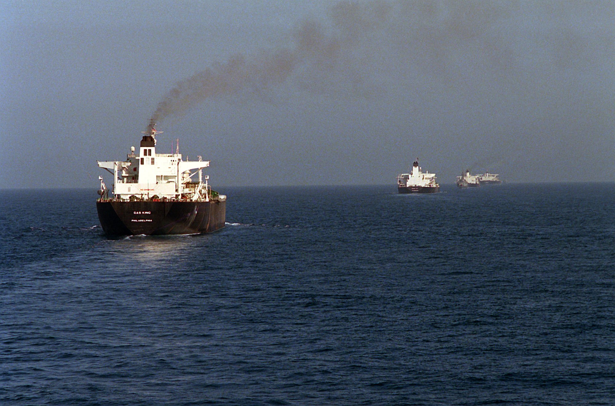 Four large tankers sail in a line across calm water, escorted by a grey US Navy warship